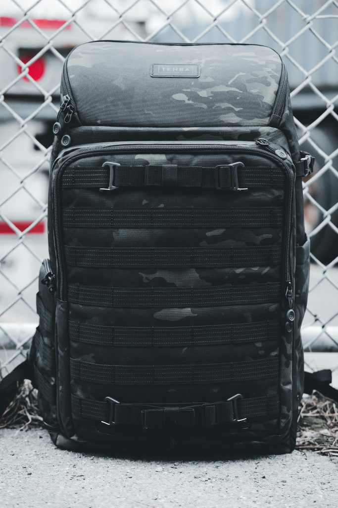 Close-up of a camouflage backpack resting on pavement against a chain link fence.