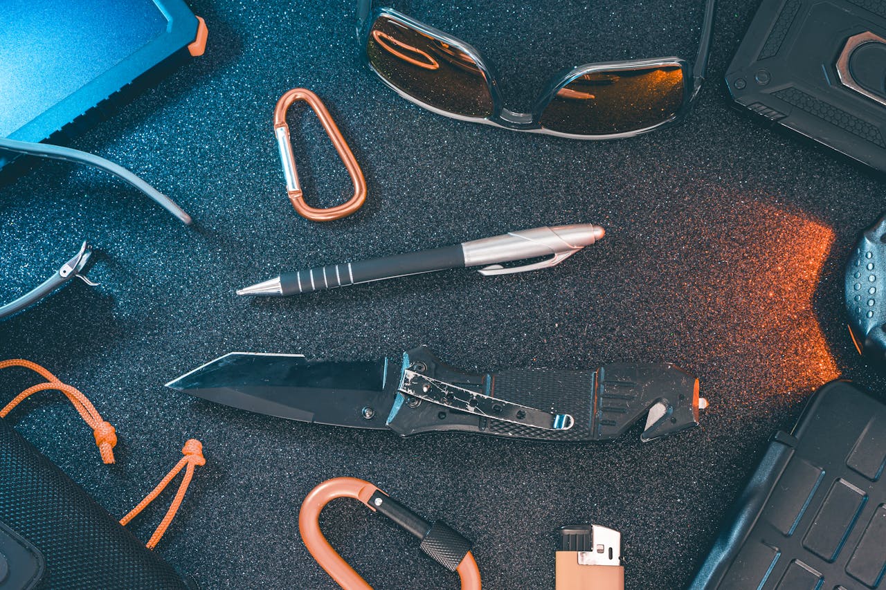 A flat lay of essential everyday carry items including a knife, pen, sunglasses, and carabiner.